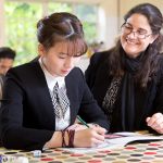 student writing in a book as a teacher looks on smiling