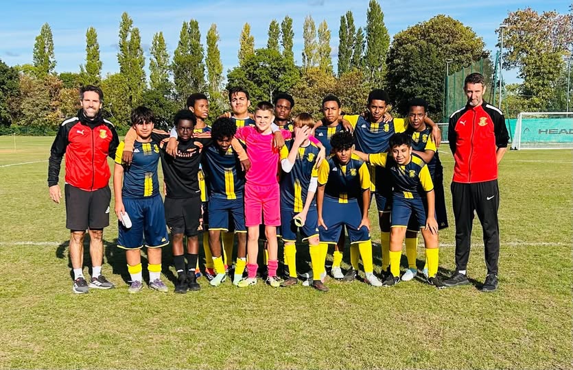 St. John’s football team in navy and yellow kit, arms linked with their two coaches in front of a goal on an artificial pitch